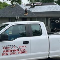 Roofing work in progress with workers atop a house and a roofing company truck parked in the foreground.