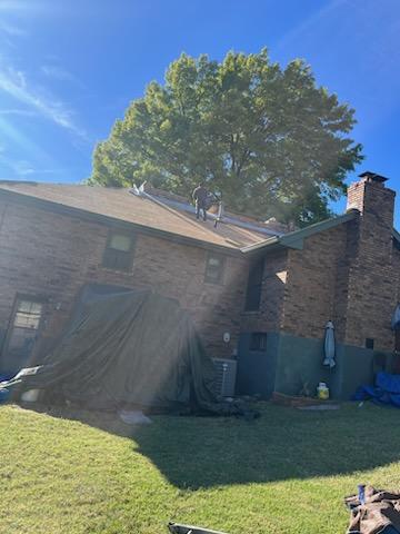 Workers on the roof of a residential building on a sunny day, with protective tarps covering the grounds and equipment nearby, likely indicating roofing renovation or repair work in progress.