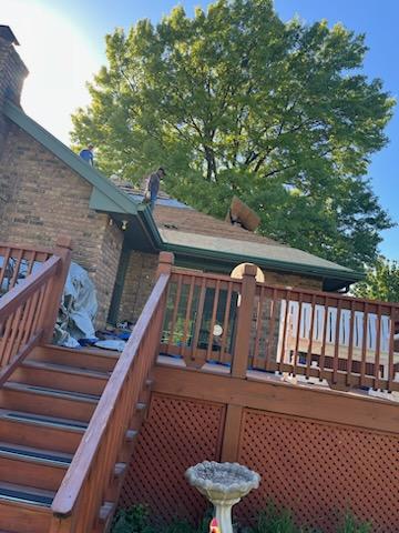 A roofer on the sloping edge of a sunlit house roof, installed by Kansas City roofers, overlooking a wooden deck with a birdbath in the foreground.